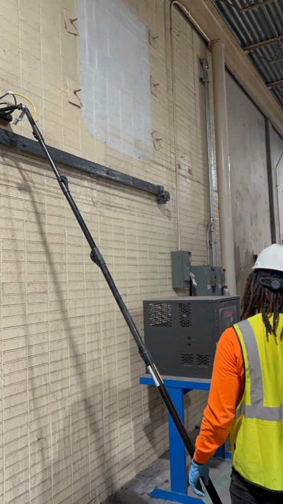 A janitor wearing a green vest using a high-rise dusting broom to clean the wall