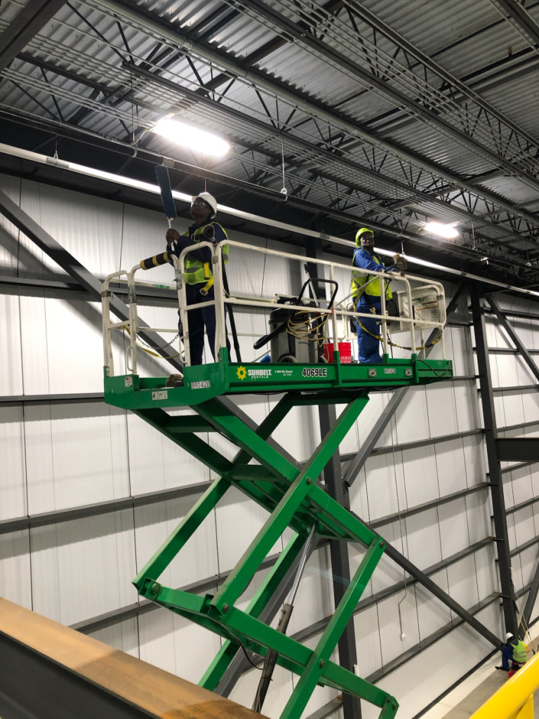 janitorial staff members on a green crane cleaning high points of the warehouse