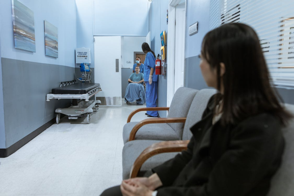 An image of a nurse pushing a patient on a wheelchair while a woman sits on a chair in a clinic