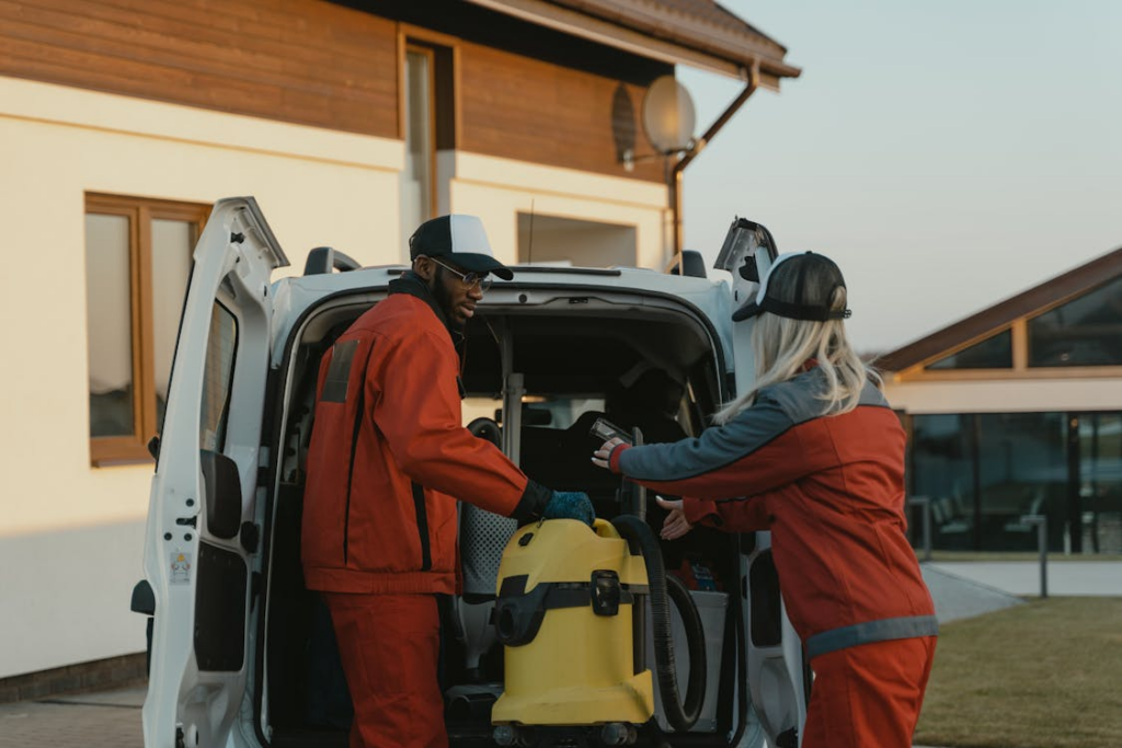 Two cleaners in orange overalls getting a vacuum cleaner from a van.